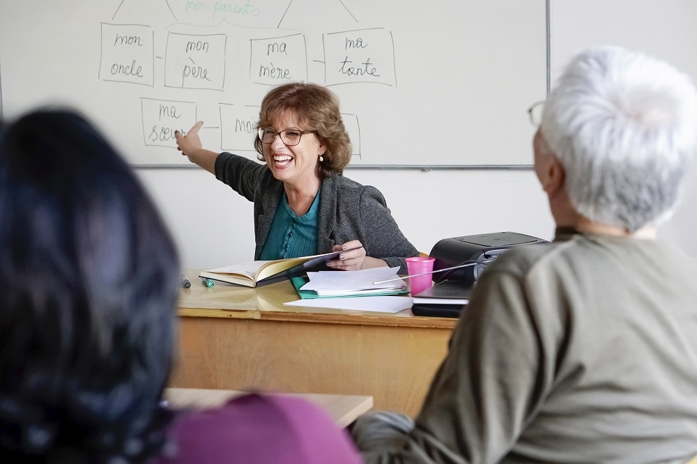 A woman leads a diverse class, engaging adults learners around a desk with French language content on the whiteboard.