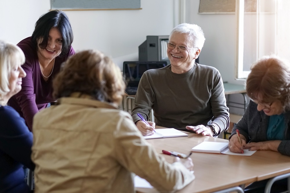 A collaborative scene featuring a diverse group of individuals at a table, while a woman focuses on writing notes or ideas. 